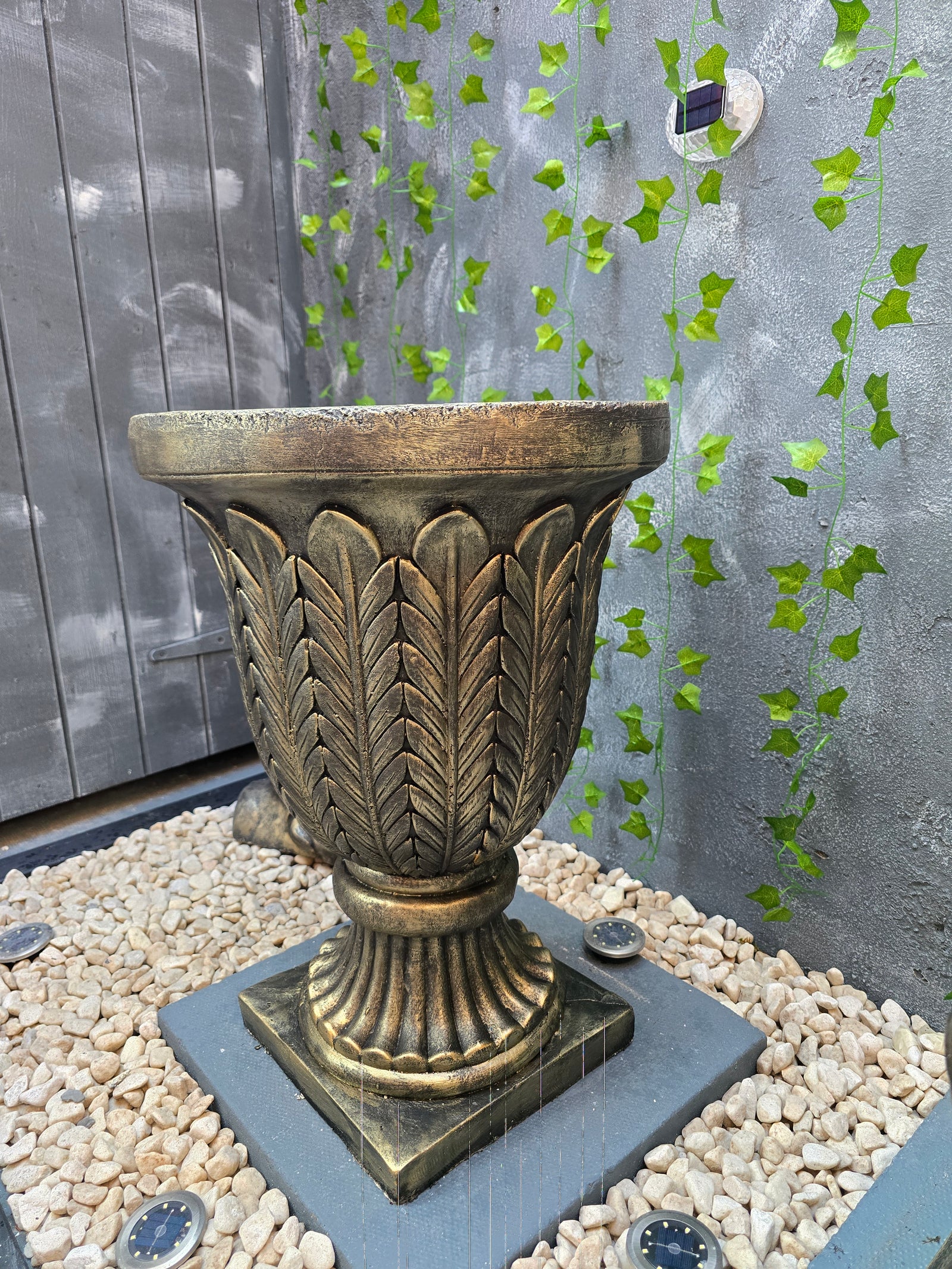 Decorative metal pedestal on a textured surface with green leaves and pebbles.