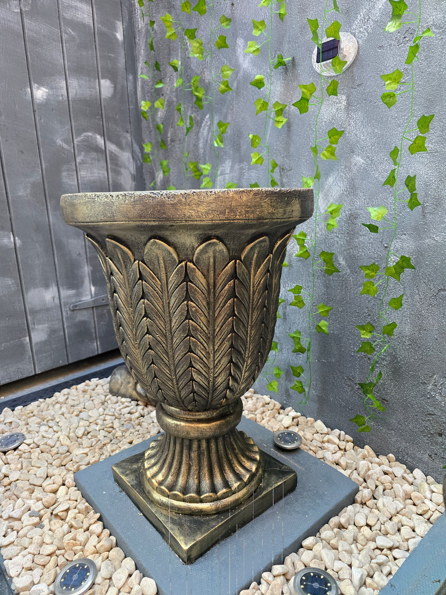Decorative metal pedestal on a textured surface with green leaves and pebbles.